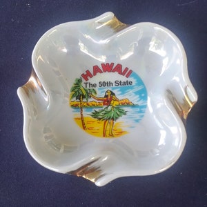 May include: White ceramic ashtray with a gold rim and a colorful image of a hula dancer on a beach with the text "HAWAII The 50th State".