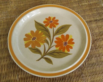 May include: A beige ceramic dinner plate with a brown rim and a floral design featuring orange, yellow, and green flowers.