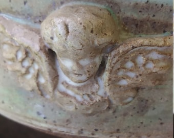 Può includere: Una ciotola in ceramica con una glassa verde e un designo di angelo scolpito. L'angelo ha le ali e un viso con gli occhi chiusi. La ciotola ha una glassa macchiata di marrone e bianco.