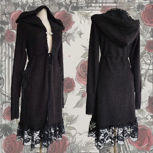 Peut inclure: Long manteau noir à capuche en tricot texturé. Le manteau présente une bordure en dentelle à l'ourlet et à la capuche, avec une fermeture à fermoir décoratif. Les manches sont longues et le manteau a une attache à la taille.