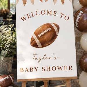 Könnte beinhalten: Ein rechteckiges, weißes Willkommensschild auf einer Holzstaffelei mit einer Aquarell-Illustration eines Footballs, braunen Wimpeln am oberen Rand und dem Text "WELCOME TO Taylor's BABY SHOWER" in brauner Schrift im Hochformat.
