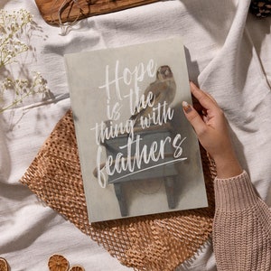 May include: A book with a white background and a bird perched on a small wooden stool. The book has the text "Hope is the thing with feathers" written in white cursive font.