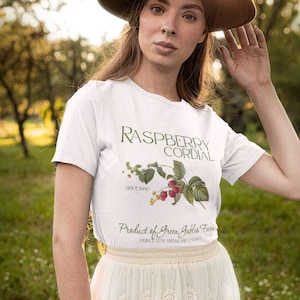 May include: A white t-shirt with a green illustration of raspberries and the text "Raspberry Cordial" and "Product of Green Gables Farm". The model is wearing a brown hat and a white tulle skirt.