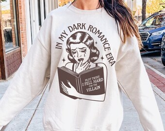 May include: A white sweatshirt with a brown graphic print of a woman reading a book. The text on the graphic says "In My Dark Romance Era, Plot Twist I Fell Hard For The Villain."