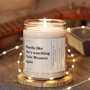 May include: A clear glass candle jar with a beige candle inside. The candle is lit and the flame is visible. The jar has a white label with the text "Smells like she's watching Little Women again" printed on it. The candle is sitting on a wooden coaster.