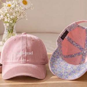 May include: Two pink baseball caps, one with "well-read" embroidered on the front. The second cap is open, revealing a floral patterned interior. A vase of daisies and books are in the background. The caps are on a wooden surface.