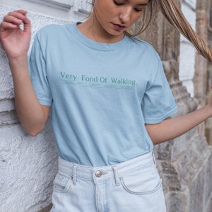May include: A woman wearing a light blue t-shirt with the text "Very Fond Of Walking" printed on it. She is wearing light blue denim shorts and has her hair pulled back in a ponytail.