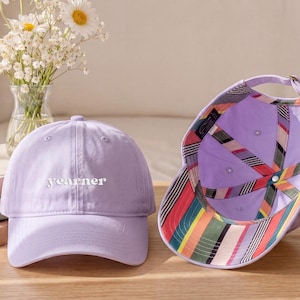 May include: Two lavender baseball caps are shown on a wooden surface. One cap is upright, with the word "yearner" in white. The other cap is turned over, revealing a colourful striped pattern on the underside. A vase of flowers and a calendar are in the background.