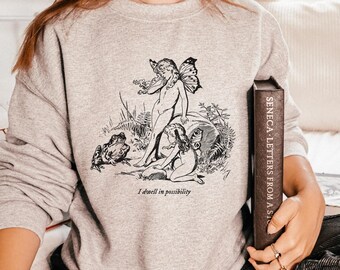 Puede incluir: Una sudadera gris con una ilustración en blanco y negro de un hada con alas y el texto "I dwell in possibility".