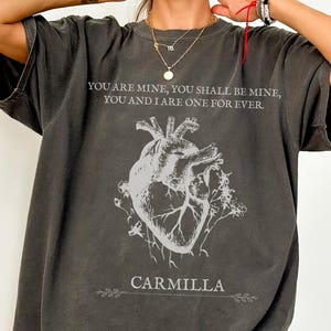 May include: Dark grey oversized t-shirt featuring a white anatomical heart design and the text "YOU ARE MINE, YOU SHALL BE MINE, YOU AND I ARE ONE FOR EVER." The word "CARMILLA" is printed below the graphic.