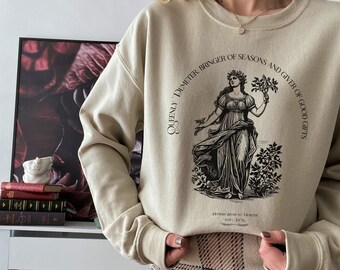 Peut inclure: Un sweat-shirt beige avec un imprimé graphique d'une femme tenant une branche de blé. Le texte sur le sweat-shirt est "Queenly Demeter, Bringer of Seasons and Giver of Good Gifts". Le sweat-shirt est associé à une jupe à carreaux.