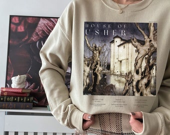 Puede incluir: Una sudadera beige con un estampado gráfico de una casa y árboles. El gráfico tiene el texto "HOUSE OF USHER" impreso en él. La sudadera la lleva puesta una persona que lleva una falda de cuadros.