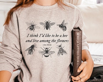 May include: A gray sweatshirt with a quote about bees and flowers printed on it. The quote reads "I think I'd like to be a bee and live among the flowers - Anne Shirley". There are five bee illustrations around the quote.
