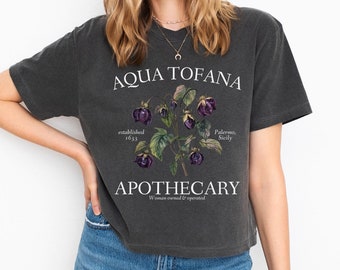 Puede incluir: Una camiseta gris oscuro con un diseño gráfico vintage que presenta una ilustración floral y el texto "Aqua Tofana Apothecary, established 1633, Palermo, Sicily, Woman owned & operated."