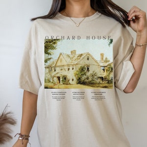 May include: A beige t-shirt with a graphic print of a house and trees. The text "ORCHARD HOUSE" is printed above the graphic. The text below the graphic reads "DO APPLE PICKING IN OUR NAMESAKE ORCHARDS", "ATTEND OUR FAMOUS AMATEUR THEATER", and "ICE SKATE IN OUR WINTER WONDERLAND".