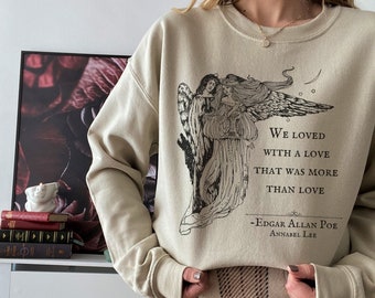 Puede incluir: Una sudadera beige con un estampado gráfico negro de dos ángeles y una cita del poema de Edgar Allan Poe "Annabel Lee": "We loved with a love that was more than love". La sudadera se combina con una falda de cuadros marrones y blancos.