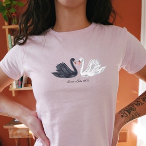 Könnte beinhalten: Ein pinkfarbenes T-Shirt mit einem schwarzen und weißen Schwanenmotiv. Der Text "Swan Lake (1875)" ist unter den Schwänen aufgedruckt.
