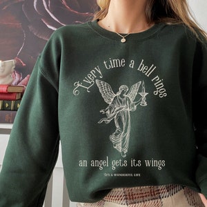 Pode incluir: Uma camisa de moletom verde escura com uma estampa gráfica branca de um anjo segurando uma vela. O texto diz "Every time a bell rings an angel gets its wings - It's a Wonderful Life".