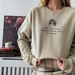 May include: A beige sweatshirt with a graphic print of a woman and the text "I wish I were a girl again, half-savage and hardy, and free. -Wuthering Heights". The sweatshirt is paired with a brown and white plaid skirt.