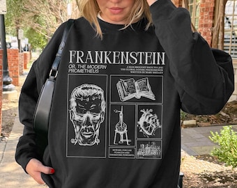 May include: Black sweatshirt featuring the word "FRANKENSTEIN" and the phrase "OR, THE MODERN PROMETHEUS" in white. The design includes illustrations of a face, a book, a medical device, and a heart. The sweatshirt is worn by a person.