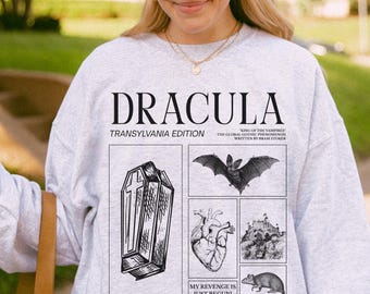May include: Light gray sweatshirt featuring the word "DRACULA" above a series of black and white illustrations. The illustrations include a coffin, bat, heart, castle, and rat. Text on the sweatshirt reads "TRANSYLVANIA EDITION" and "MY REVENGE IS JUST BEGUN!"