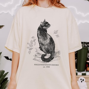Op de afbeelding: Cr&egrave;mekleurig T-shirt met een illustratie van een zwarte kat die op boeken zit, met de tekst "Halloweentown Books est. 1998". Het ontwerp bevat bladeren, wat een griezelig, boekachtig thema cre&euml;ert. Het shirt heeft korte mouwen.