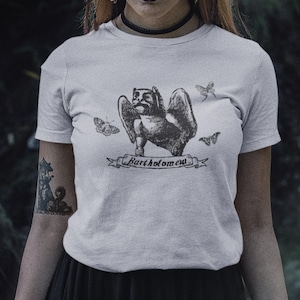 Könnte beinhalten: Ein hellgraues Kurzarm-T-Shirt mit einem dunkelgrauen Grafikdruck einer geflügelten, gargoyleartigen Kreatur, die von drei kleinen Motten umgeben ist. Unter der Kreatur befindet sich ein Banner mit dem Text Bartholomew in Serifenschrift.