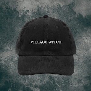 Puede incluir: Una gorra de béisbol de pana negra con el texto "VILLAGE WITCH" bordado en blanco en la parte delantera.