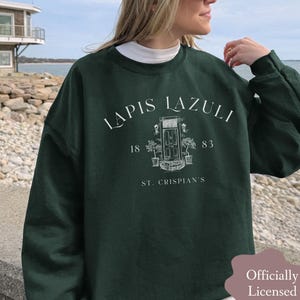 以下が含まれることがあります： ダークフォレストグリーンのクルーネックトレーナー。LAPIS LAZULI、1883、ST. CRISPIAN'Sの文字と、ドアと鉢植えの線画が白でプリントされています。茶色のバッジにはOfficially Licensed Collection!と記載されています。