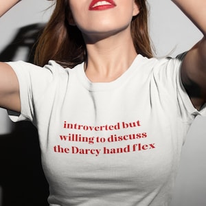 Pode incluir: Camiseta branca com o texto vermelho: "introverted but willing to discuss the Darcy hand flex". A camiseta é de gola redonda.