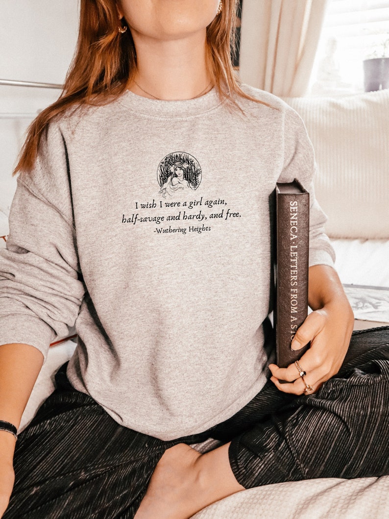 May include: A gray sweatshirt with a quote from Wuthering Heights: "I wish I were a girl again, half-savage and hardy, and free." The quote is printed in black text and is surrounded by a small illustration of a woman in a field.