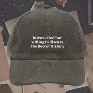 May include: A green corduroy baseball cap with white embroidered text that reads "introverted but willing to discuss The Secret History".