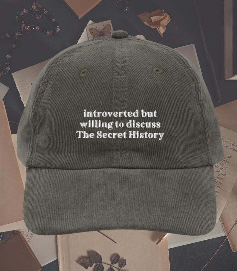 May include: A green corduroy baseball cap with white embroidered text that reads "introverted but willing to discuss The Secret History".
