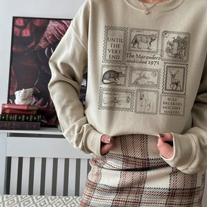 K&ouml;nnte beinhalten: Ein beiger Pullover mit einem grafischen Druck von Briefmarken mit verschiedenen Illustrationen, darunter ein Wolf, ein Schloss, eine Ratte, ein Hirsch, ein Teleskop und ein Hund. Der Text auf den Briefmarken lautet "Until the very end", "The Marauders established 1971" und "Rule Breakers Mischief Makers".