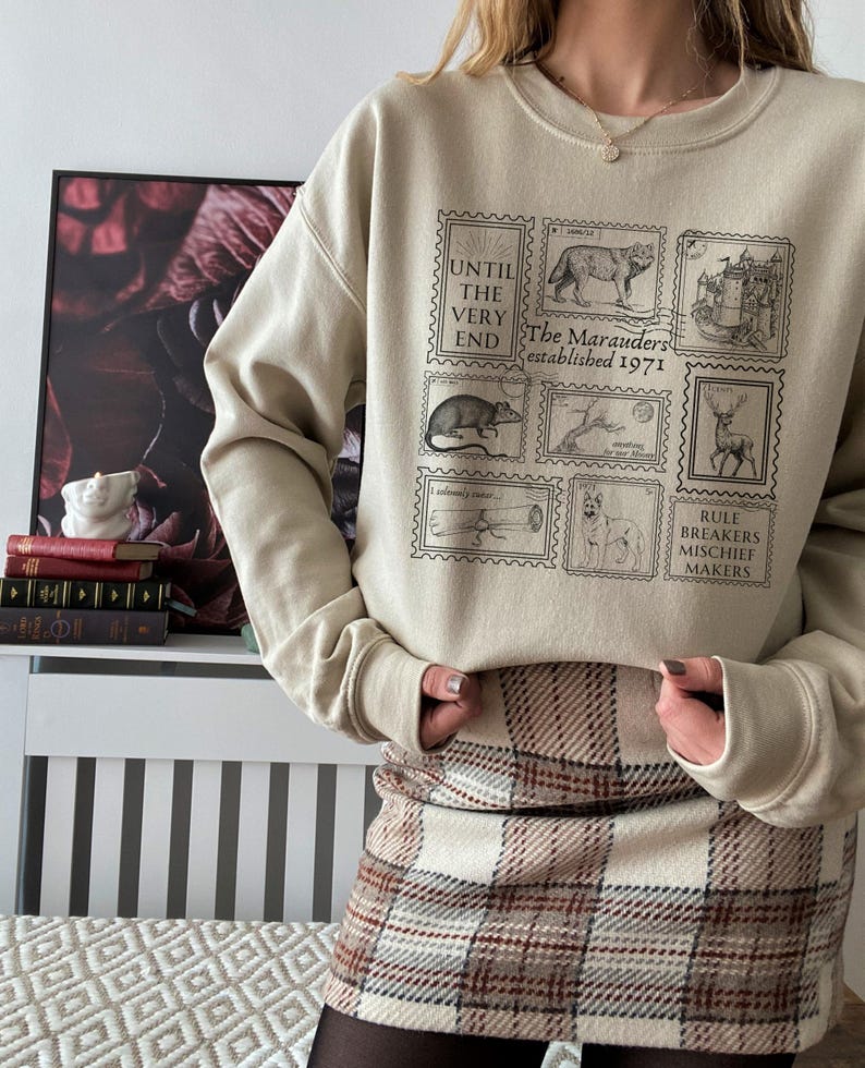 K&ouml;nnte beinhalten: Ein beiger Pullover mit einem grafischen Druck von Briefmarken mit verschiedenen Illustrationen, darunter ein Wolf, ein Schloss, eine Ratte, ein Hirsch, ein Teleskop und ein Hund. Der Text auf den Briefmarken lautet "Until the very end", "The Marauders established 1971" und "Rule Breakers Mischief Makers".