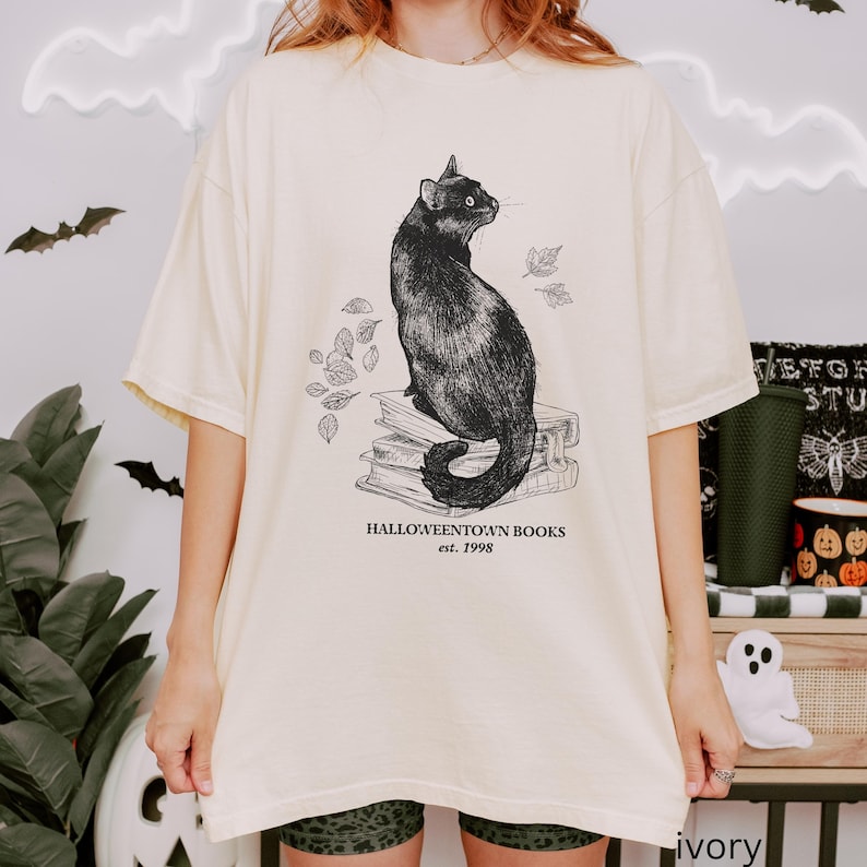 Op de afbeelding: Cr&egrave;mekleurig T-shirt met een illustratie van een zwarte kat die op boeken zit, met de tekst "Halloweentown Books est. 1998". Het ontwerp bevat bladeren, wat een griezelig, boekachtig thema cre&euml;ert. Het shirt heeft korte mouwen.