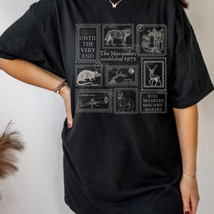 May include: Black t-shirt with a white graphic print of a series of stamps featuring images of a wolf, a castle, a rat, a tree, a deer, a wand, and a dog. The stamps have text that reads "Until the very end", "The Marauders established 1971", "I solemnly swear...", and "Rule Breakers Mischief Makers".