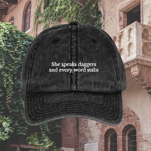 Op de afbeelding: Zwarte denim baseballpet met witte geborduurde tekst die "She speaks daggers and every word stabs" luidt.