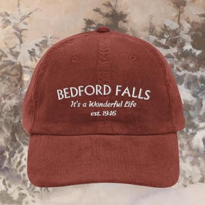 Pode incluir: Boné de beisebol de veludo cotelê bordô com o texto "BEDFORD FALLS" e "It's a Wonderful Life est. 1946" bordado em branco. O boné tem uma aba curva e um topo arredondado. O fundo é uma pintura desfocada de uma cena de inverno.