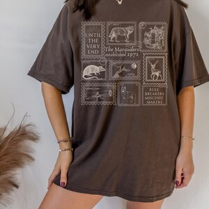 May include: A brown oversized t-shirt with a graphic print of vintage postage stamps featuring illustrations of animals and text that reads "Until the very end, The Marauders established 1971, Rule Breakers Mischief Makers".