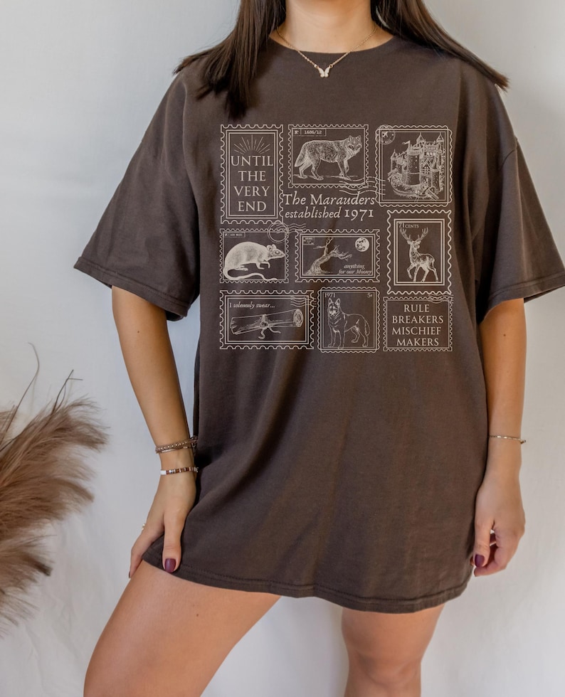 May include: A brown oversized t-shirt with a graphic print of vintage postage stamps featuring illustrations of animals and text that reads "Until the very end, The Marauders established 1971, Rule Breakers Mischief Makers".