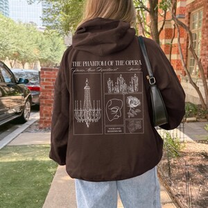 May include: A brown hoodie with a hood and a graphic design on the back. The design includes text that reads "THE PHANTOM OF THE OPERA" and various illustrations, including a chandelier, a mask, and a rose. The person is wearing light blue jeans.