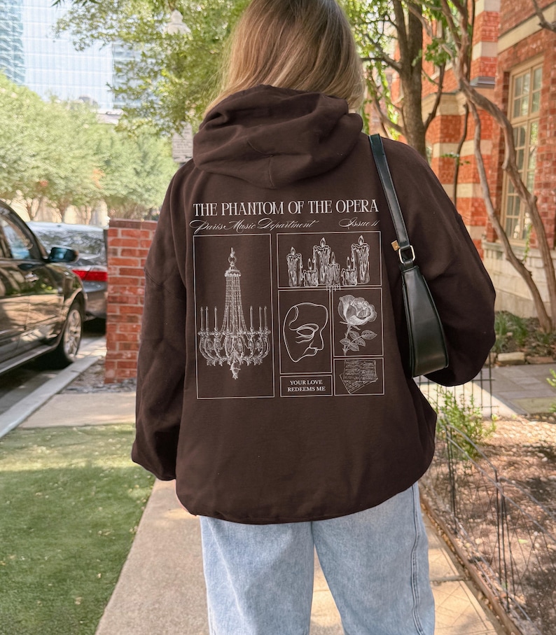 May include: A brown hoodie with a hood and a graphic design on the back. The design includes text that reads "THE PHANTOM OF THE OPERA" and various illustrations, including a chandelier, a mask, and a rose. The person is wearing light blue jeans.