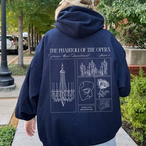 May include: Navy blue hoodie with a white graphic design featuring elements from "The Phantom of the Opera." The design includes a chandelier, candles, a mask, a rose, and the text "YOUR LOVE REDEEMS ME."