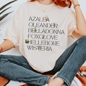 May include: A beige t-shirt with the words "Azalea", "Oleander", "Belladonna", "Foxglove", "Hellebore", and "Wisteria" printed on it. Each word is decorated with a small floral illustration.