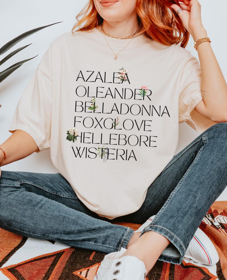 May include: A beige t-shirt with the words "Azalea", "Oleander", "Belladonna", "Foxglove", "Hellebore", and "Wisteria" printed on it. Each word is decorated with a small floral illustration.