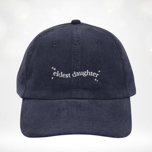 May include: Navy blue corduroy baseball cap featuring the embroidered words "eldest daughter" in white. The hat has a curved peak and a button on top. The text is framed by small star motifs.