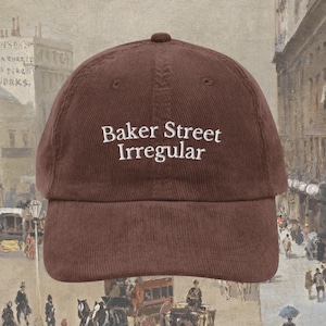 Pode incluir: Boné de beisebol de veludo cotelê marrom com o texto "Baker Street Irregular" em branco. O boné tem uma aba curva e um topo arredondado. O fundo é uma pintura em estilo vintage de uma cena de rua.