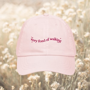 May include: A light pink baseball cap with the embroidered text "very fond of walking" in a darker pink script. The cap has a curved brim and a classic six-panel design, perfect for outdoor activities.