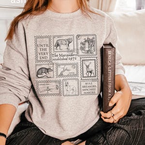 K&ouml;nnte beinhalten: Ein graues Sweatshirt mit einem grafischen Druck von Briefmarken mit verschiedenen Bildern, darunter ein Wolf, ein Schloss, eine Ratte, ein Baum, ein Hirsch, eine Schriftrolle und ein Hund. Der Text auf den Briefmarken lautet "Until the very end", "The Marauders established 1971" und "Rule Breakers Mischief Makers".